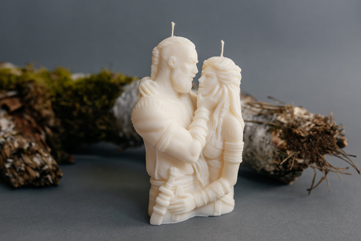 An off-white sculpted candle shaped like a romantic couple embracing, displayed on a dark gray surface with mossy logs in the background.