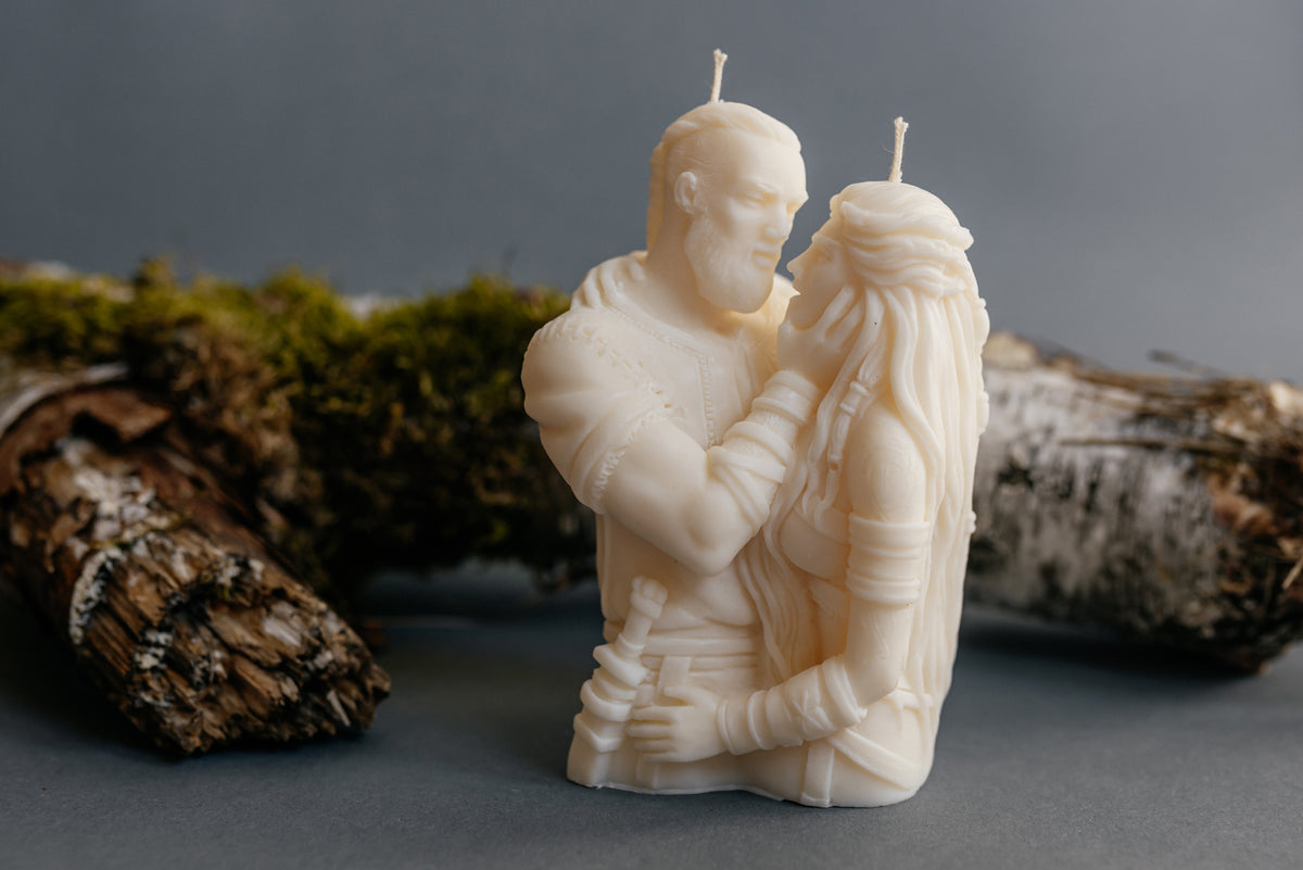 An off-white carved candle shaped like a couple embracing, displayed on a dark gray surface with mossy logs in the background.