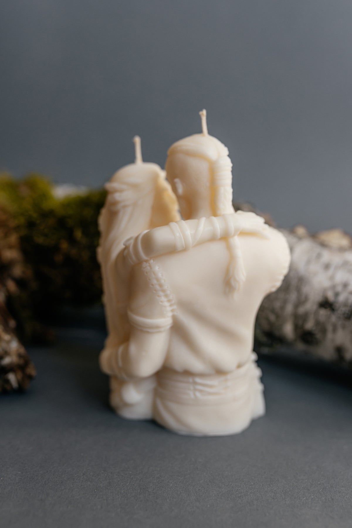An off-white sculptural candle shaped like two embracing figures, photographed on a dark gray surface with blurred natural props in the background.