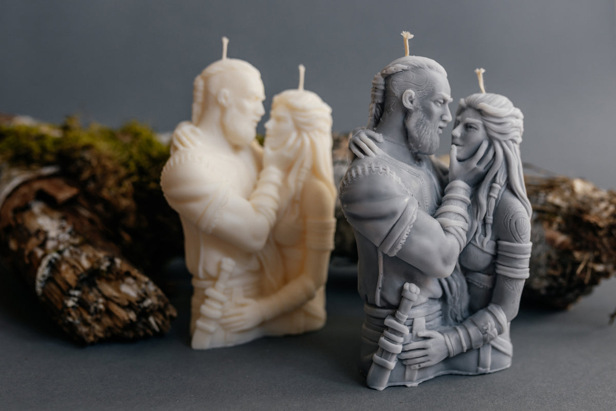Two sculpted pillar candles shaped as embracing figures, shown in off-white and matte gray on a dark gray surface with rustic wood props in the background.