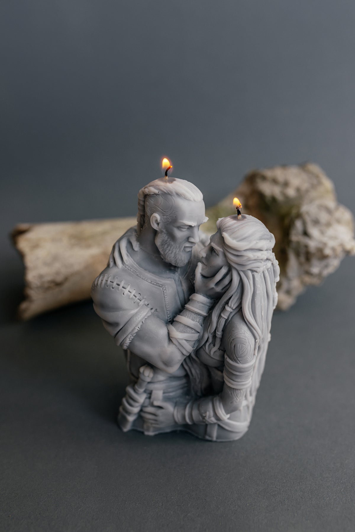 A gray sculpted candle shaped like a embracing couple, with two small lit wicks on top of the heads and shoulders. The candle is photographed on a dark gray surface with a blurred driftwood log in the background.