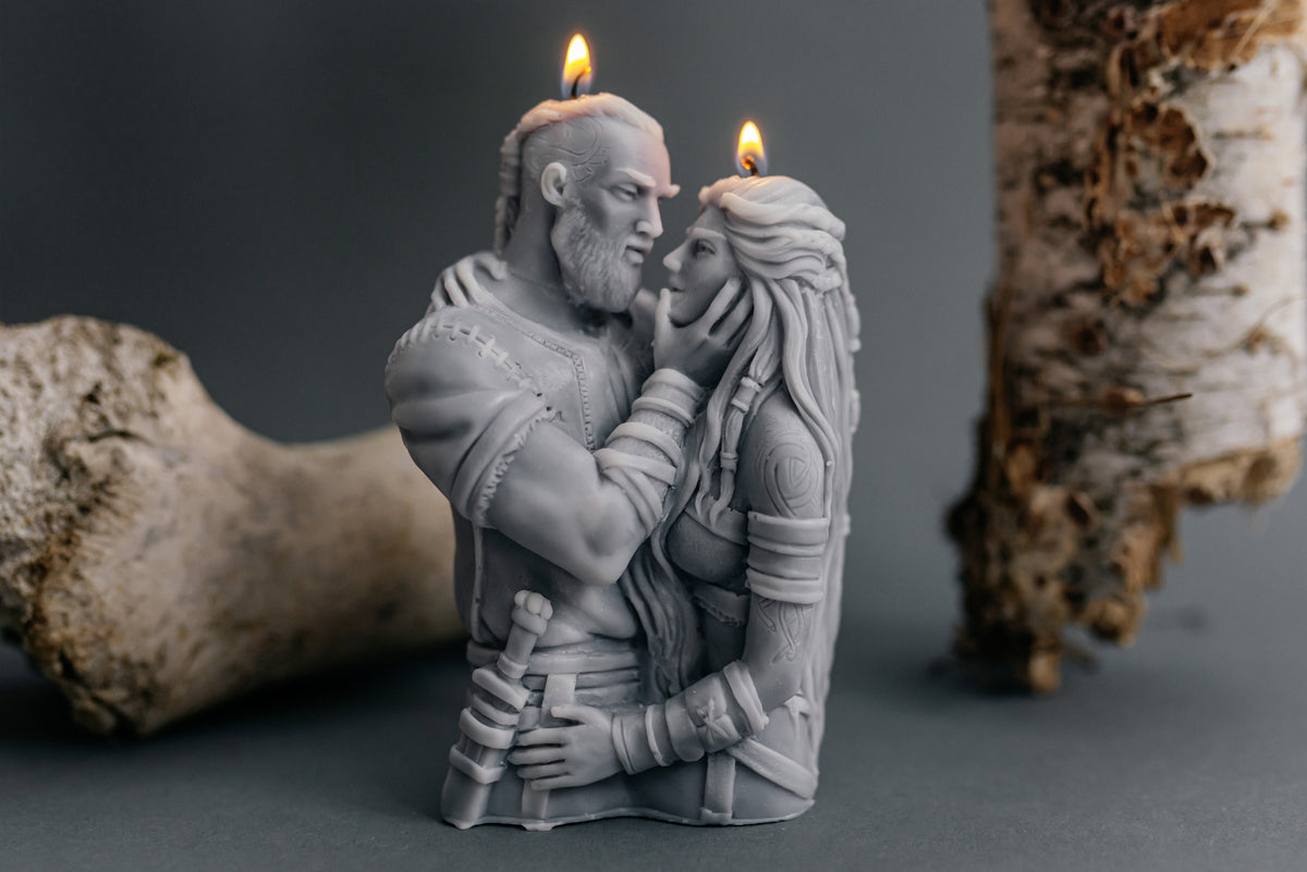 A gray sculpted candle shaped like a embracing couple, with two lit wicks, displayed on a dark surface with driftwood props in the background.