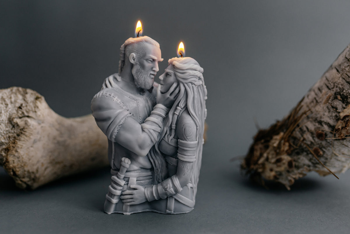 A gray sculpted candle shaped like a couple embracing, with two lit candles on their heads, photographed against a dark gray background with driftwood props on both sides.
