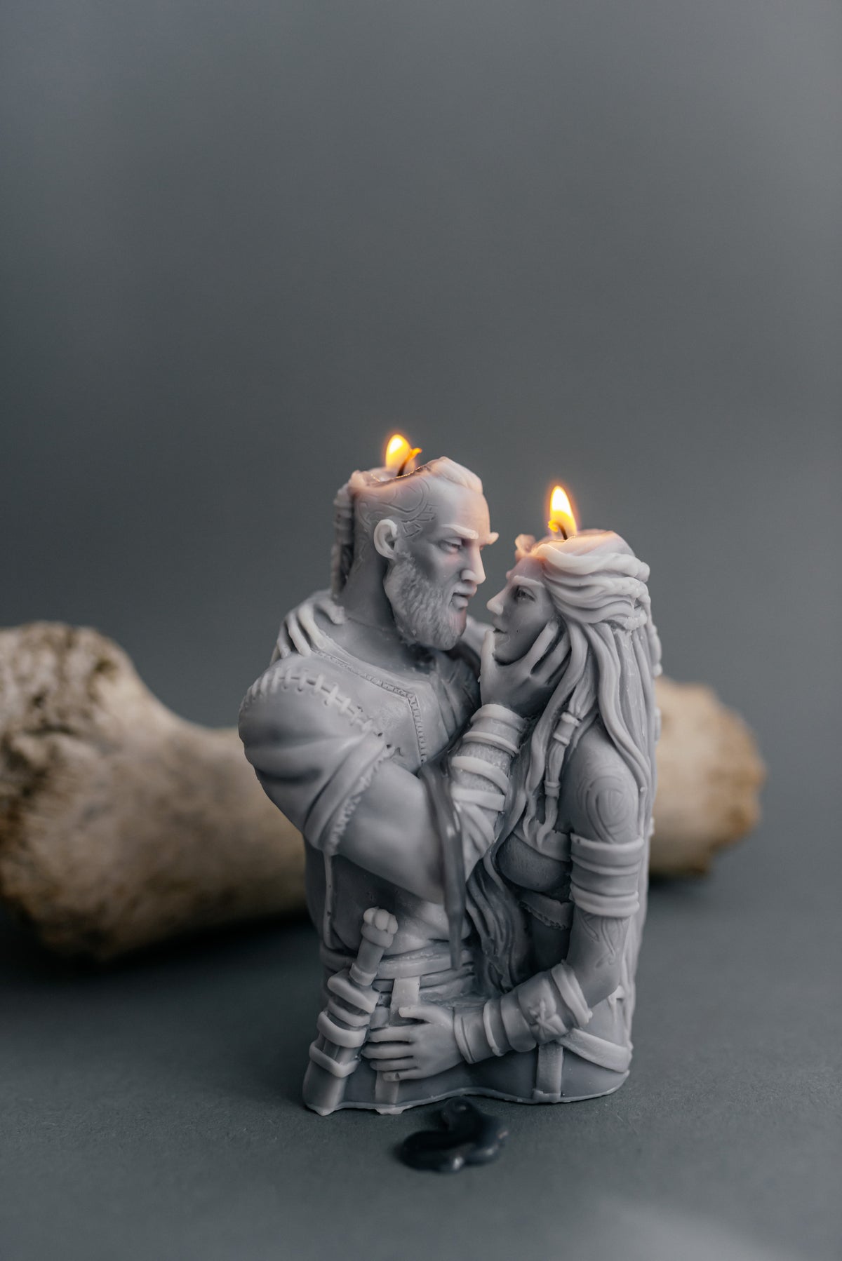 A gray sculpted candle shaped like a bearded man embracing a woman, with two small lit candles inserted into their heads. The candle is photographed on a dark gray surface with a blurred bone-like prop in the background.