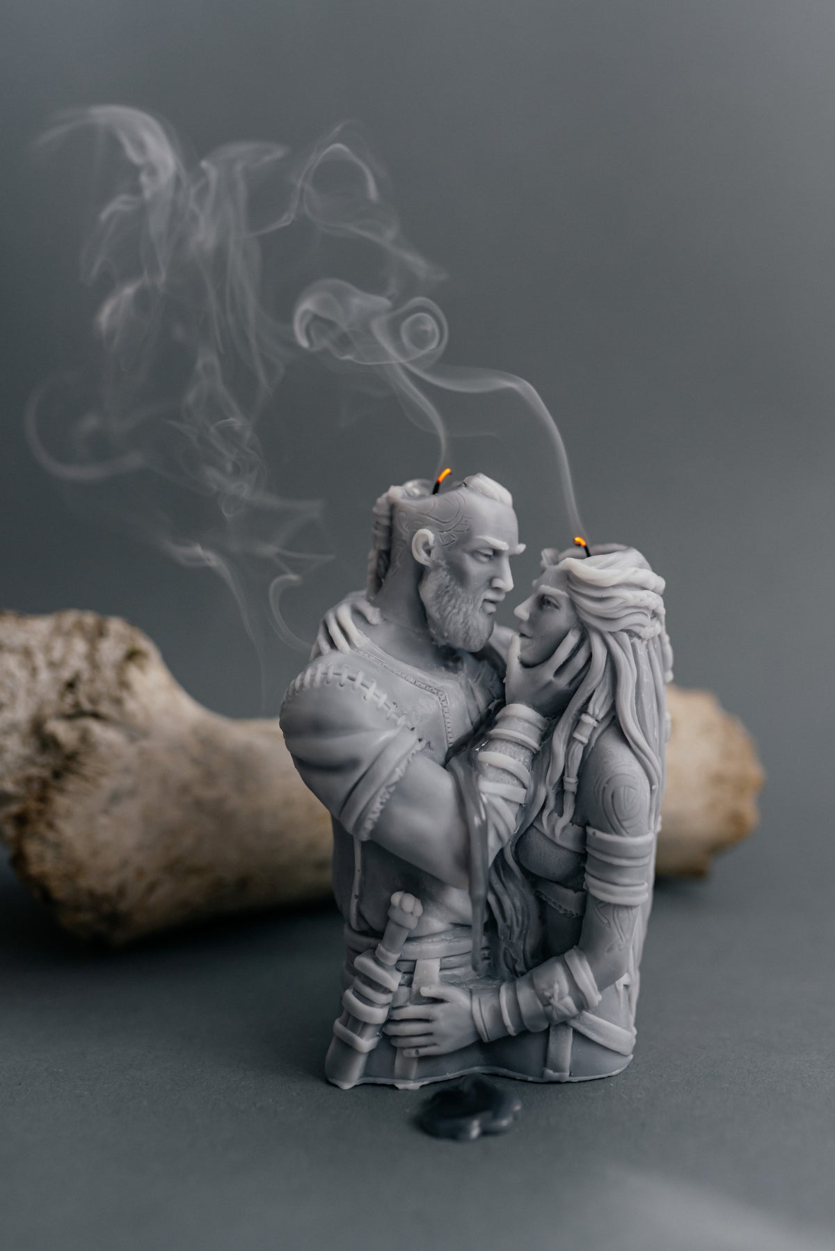A gray resin figurine of a bearded warrior embracing a woman, with smoke rising from their mouths like incense sticks, set against a dark gray background with a blurred bone prop.