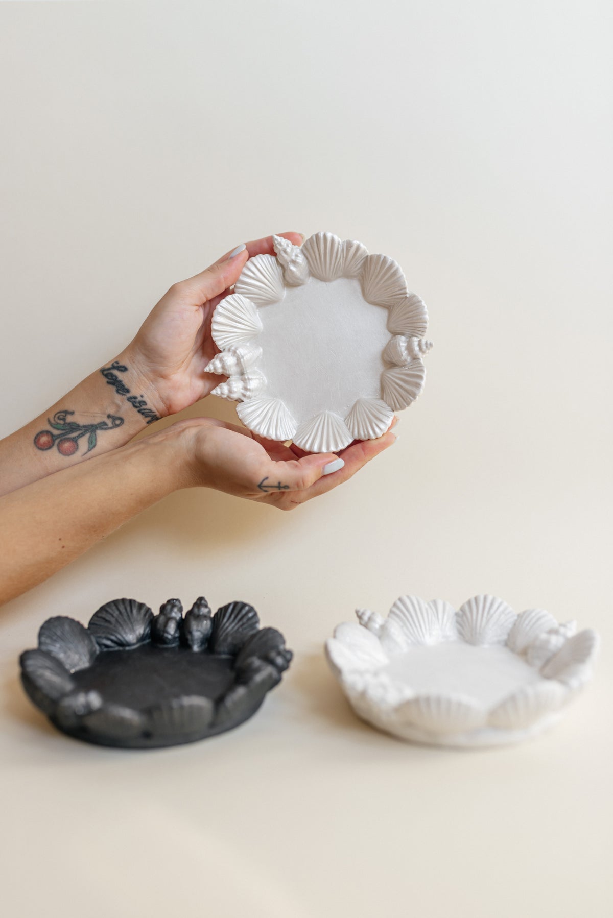 A hand holds a small scalloped plaster dish in warm off-white, with two matching dishes below on a cream background—one in dark charcoal and one in off-white.