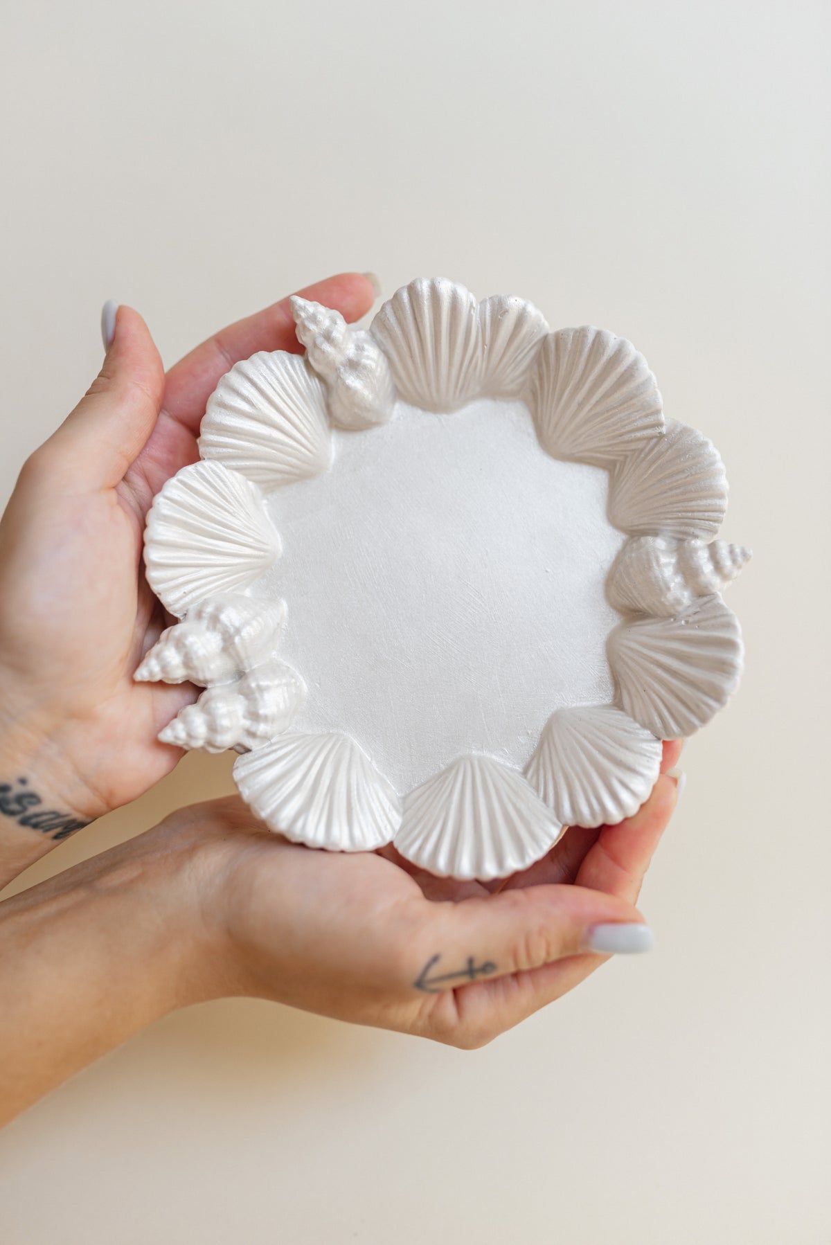 A pair of hands holding a round white plaster tray decorated with seashell and seashell-like raised patterns around the rim, against a plain light beige background.
