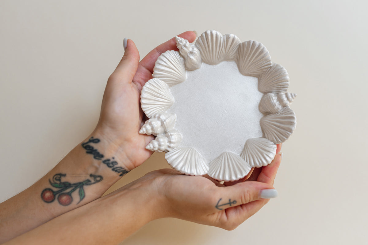 A person holds a round white plaster tray decorated with a border of seashells and small seashell motifs, set against a plain beige background.