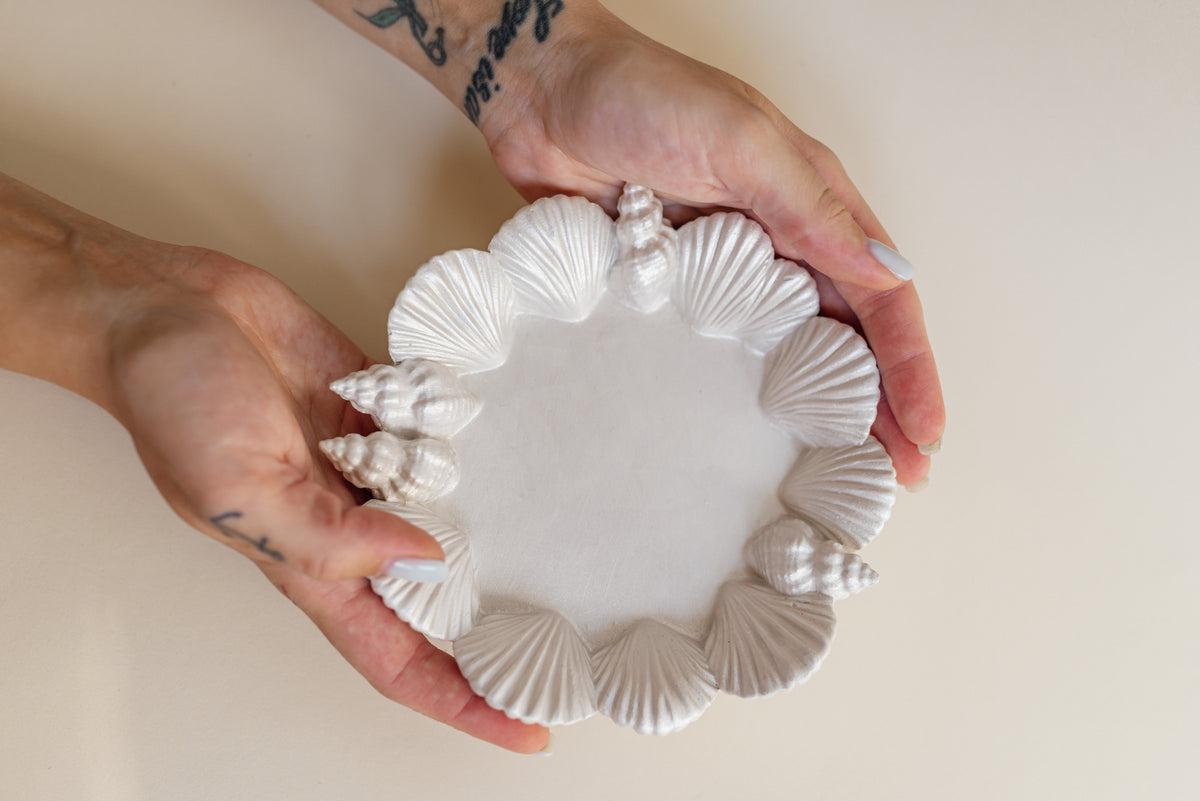 A person's hands holding a round white plaster  plate decorated with a border of realistic seashells.