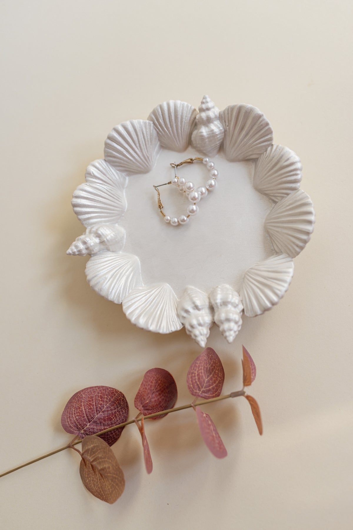 White plaster shell-shaped tray with seashell and shell-like sculptural details, styled with pearl earrings and dried pinkish-brown leaves on a warm cream background.