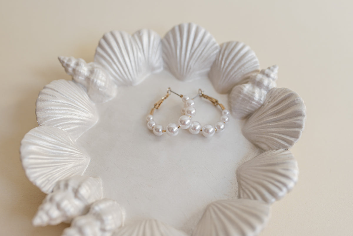 White pearl hoop earrings with gold-tone wire hangers, displayed on a white ceramic dish decorated with seashells against a warm beige background.