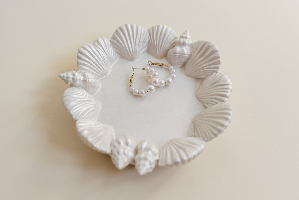 White seashell-shaped jewelry stand holding a pearl hoop earring and a gold beaded necklace, photographed on a warm cream background.