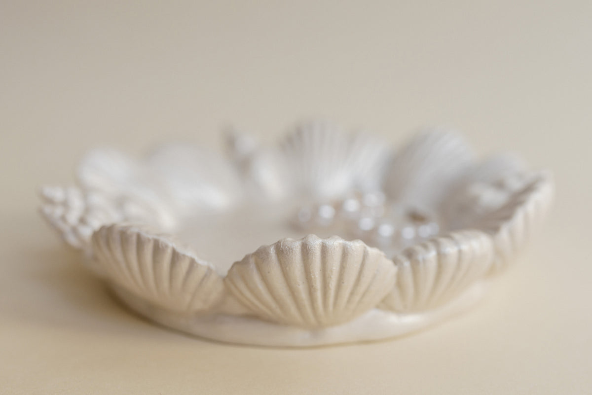 A white ceramic dish with a scallop shell design, holding a small cluster of white pearls, set against a soft beige background.