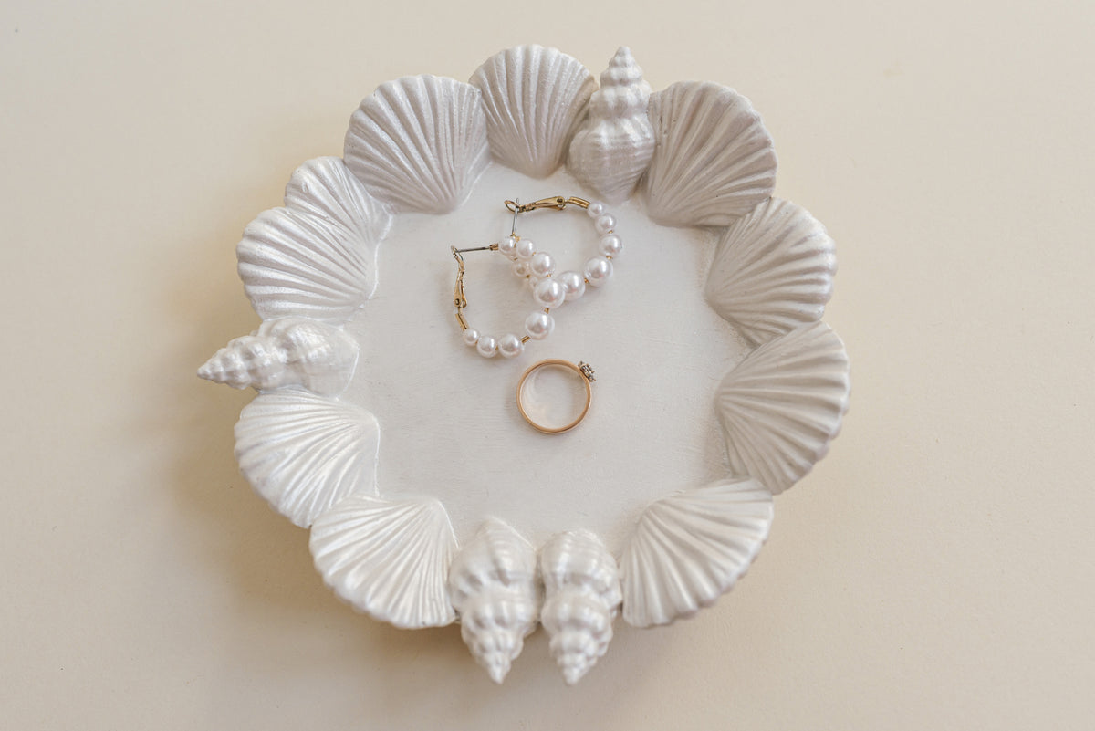 A white plaster jewelry tray designed with seashell patterns around the rim, holding a pearl hoop earring and a gold ring on a light beige background.