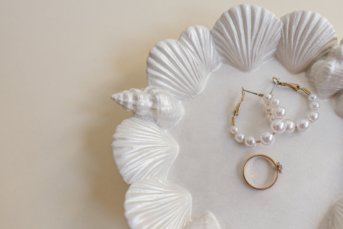 White pearl jewelry set including hoop earrings and a ring, displayed on a white shell-shaped tray against a beige background.