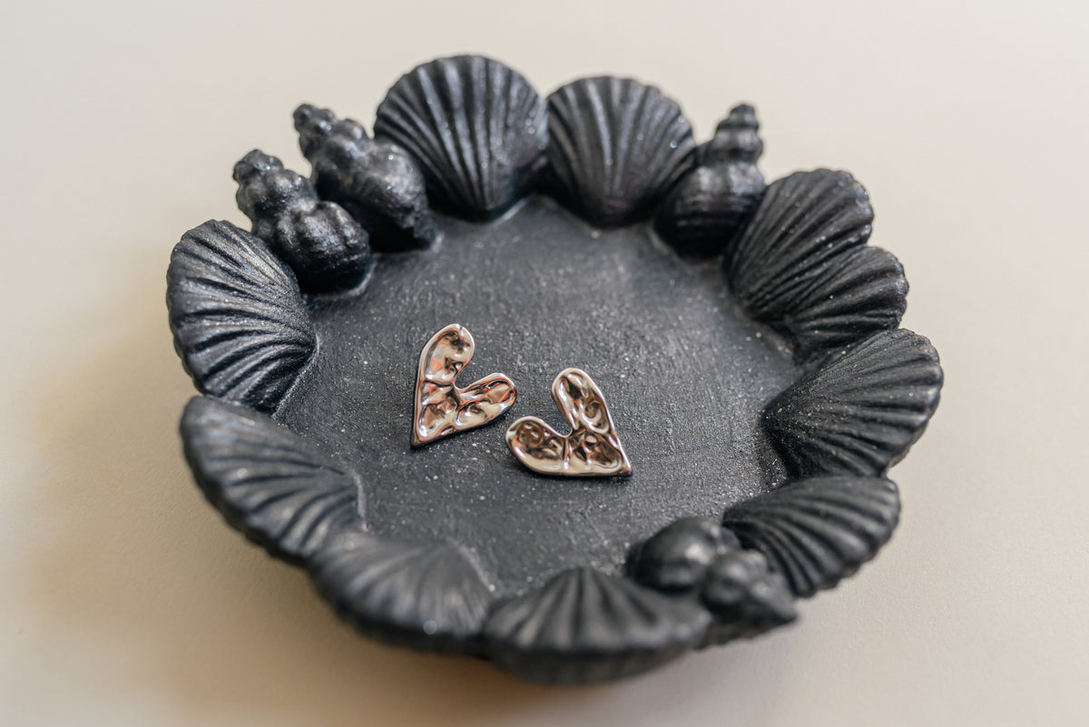 A pair of textured metallic heart earrings in a rose gold tone, displayed inside a dark gray shell-shaped dish on a light beige background.