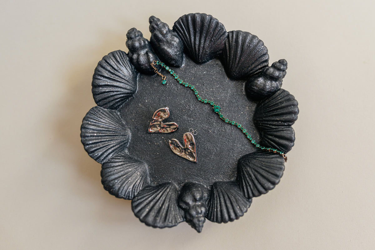 A round black jewelry tray with a scallop shell design around the edge, holding three small gold-toned heart-shaped charms and a green beaded strand, photographed against a plain light beige background.