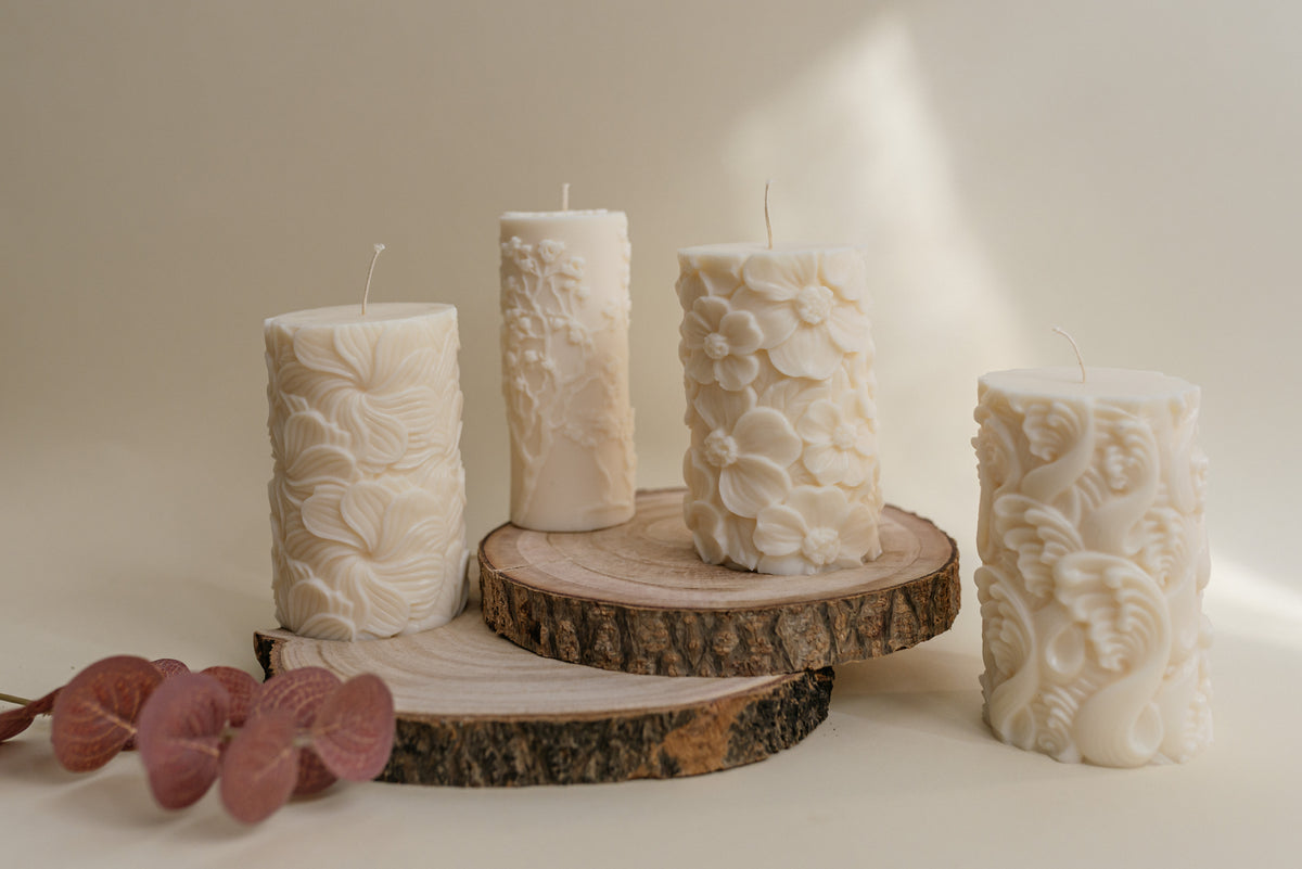 Four cream-colored pillar candles with embossed floral and leaf patterns, styled on rustic wood slices with dried pink flowers against a warm beige background.