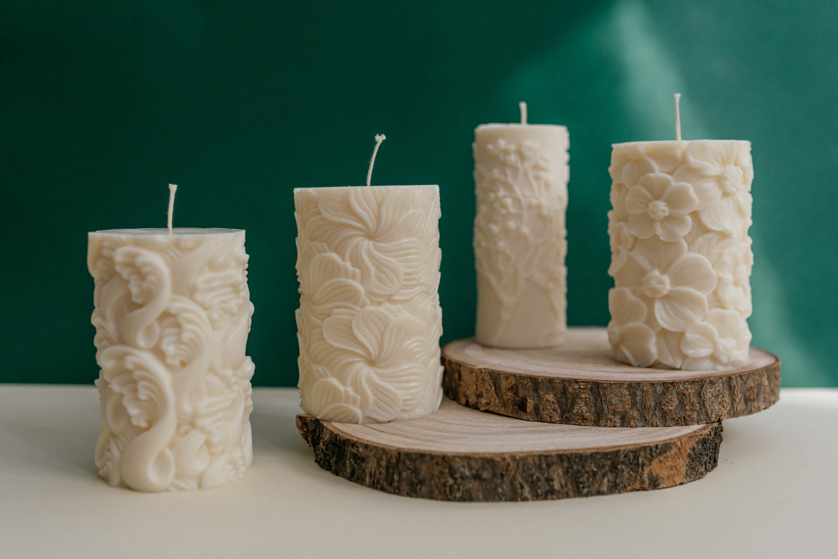 Four cream-colored pillar candles with embossed floral and scroll patterns, displayed on rustic wood slices against a dark green background.