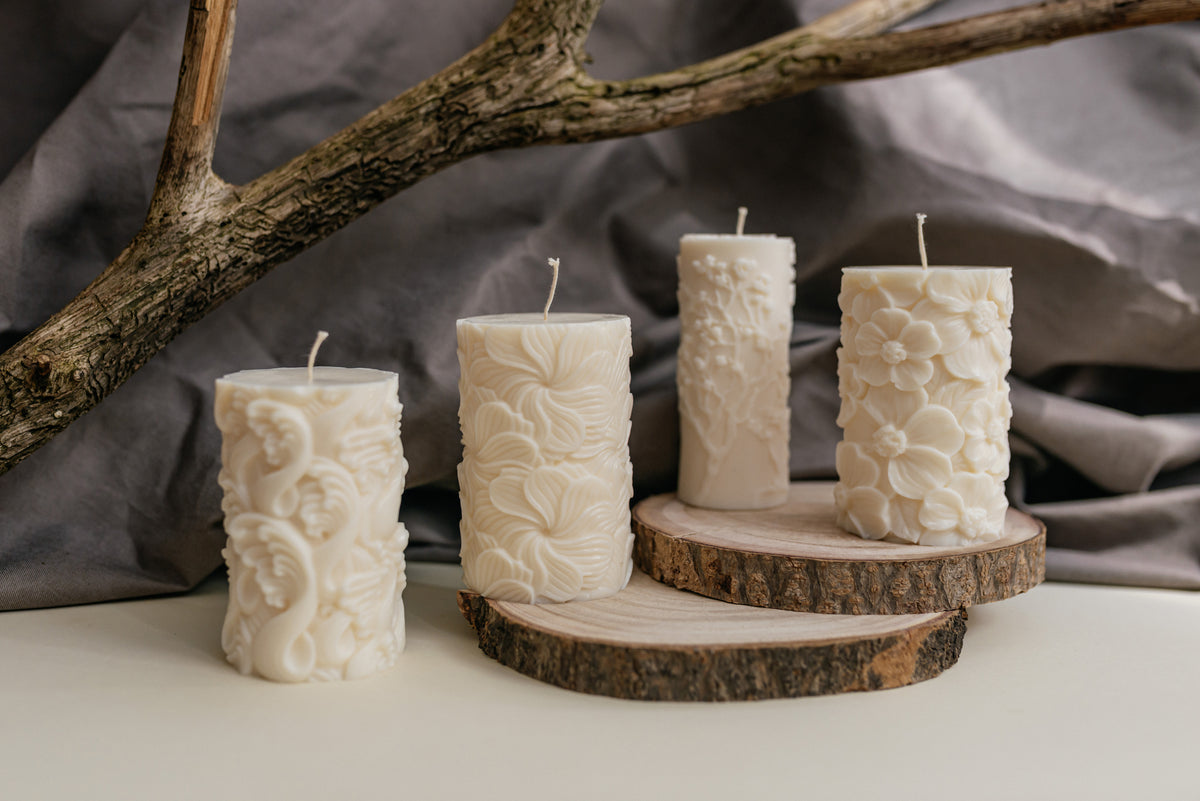 Four white wax candles with embossed floral and scroll patterns, displayed on rustic wood slices against a draped gray fabric background with a branch prop.